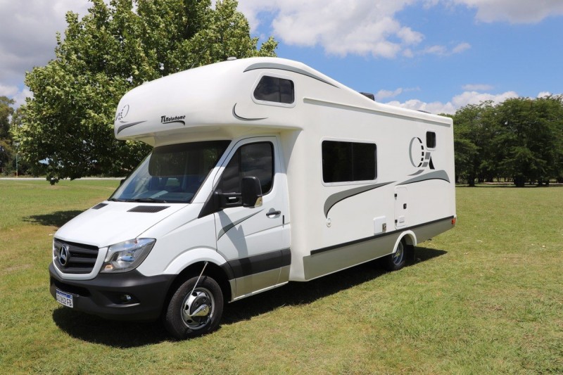 A white Mercedes motorhome parked on a grassy field with trees and a blue sky in the background, featuring large windows and streamlined design accents.